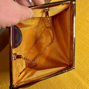 Puede incluir: Un bolso de mano estilo vintage con marco de metal dorado y apertura de bisagra. El interior está forrado con una tela de color amarillo mostaza vibrante y cuenta con una fina cadena de metal dorado. Se observa una pequeña etiqueta de cuero.
