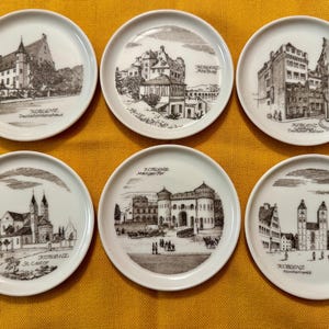 Könnte beinhalten: Set aus sechs weißen Keramikplatten, jede mit einer Schwarz-Weiß-Illustration eines Gebäudes oder Stadtbildes. Der Name des Ortes ist unter dem Bild aufgedruckt. Die Teller sind auf einer gelben Oberfläche angeordnet.