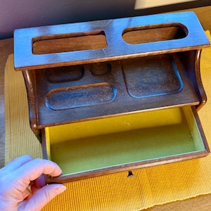 May include: A dark brown wooden desk organizer featuring a top tier with two rectangular slots and a middle section with four recessed compartments of varying shapes. The unit includes a single pull-out drawer lined with a mustard yellow felt interior.
