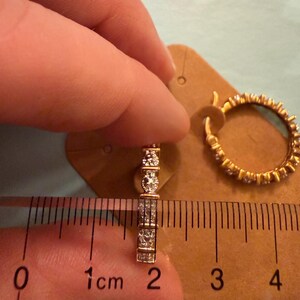 May include: A pair of gold hoop earrings featuring a row of round clear stones. One earring is positioned against a ruler showing a width of approximately 0.75 inches. The earrings are mounted on a brown cardboard display card.
