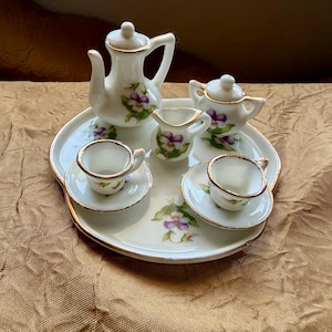 May include: A miniature white porcelain tea set with gold trim, featuring a teapot, sugar bowl, creamer, and two teacups with saucers. The set is decorated with purple floral designs and rests on a matching tray.
