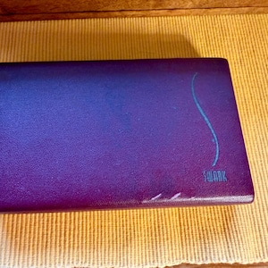 May include: A rectangular, deep purple storage case with a textured, leather-like surface. The case features a thin, light blue vertical wavy line on the right side and the word SWANK printed in light blue lettering near the bottom right corner.