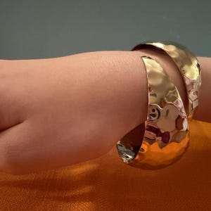 May include: A gold-toned cuff bracelet with a wide, open-ended design and a hammered, reflective surface. The bracelet features two tapered, overlapping ends that curve around the wrist, creating a bold, metallic accessory with a textured finish.