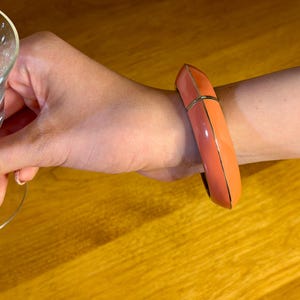 May include: A coral and orange bangle bracelet with gold-colored trim. The bracelet is worn on a wrist, and a clear glass with a floral design is held in the hand.