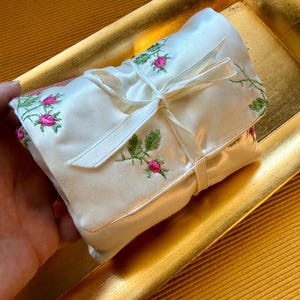 Pode incluir: Um rolo de tecido de cetim branco com botões de rosa cor-de-rosa bordados, caules e folhas verdes. O tecido é preso com uma fita de cetim branco a condizer atada num laço, criando uma textura macia e suave com detalhes florais delicados.