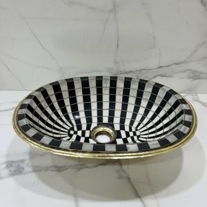 May include: An oval-shaped vessel sink featuring a repeating black and white chequered pattern across its interior surface. The basin is finished with a metallic gold-toned rim that encircles the top edge and a central circular drain opening.