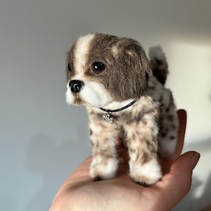 May include: A plush toy dog with a brown and white spotted coat, floppy ears, and a black collar with a small charm. The dog has a white muzzle and dark eyes, giving it a lifelike appearance. The toy is held in a hand.
