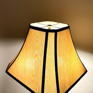 May include: A bell-shaped lamp shade with a light tan, textured fabric surface. The shade features a distinct octagonal silhouette with sharp, dark black trim outlining each of the eight panels, creating a structured and geometric appearance.