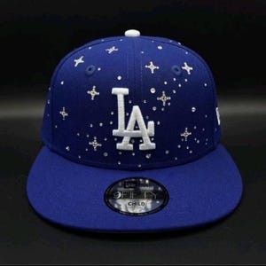 May include: A royal blue baseball cap with a flat brim. The cap features the white letters "LA" on the front, embellished with sparkling accents. The cap has a white button on top and a "9FIFTY CHILD" sticker on the brim.