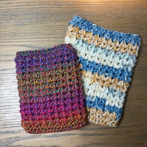 May include: Two handmade crochet cozies. One is a vibrant mix of red, orange, yellow, green, and purple. The other features blue, cream, and gold horizontal stripes. Both are made with a textured stitch.