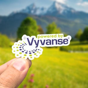 May include: A white die-cut sticker featuring the text "powered by Vyvanse" in dark purple, accompanied by a circular graphic pattern composed of small purple and lime green dots arranged in a swirling, radiating design on a white background.