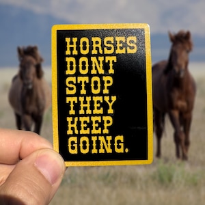 Puede incluir: Una pegatina rectangular con el texto "HORSES DONT STOP THEY KEEP GOING" en letras amarillas sobre un fondo negro. La pegatina se sostiene frente a un fondo borroso con dos caballos marrones en un campo.