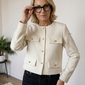 Op de afbeelding: Crèmekleurig, op maat gemaakte jas met goudkleurige knopen en zakdetails. De jas heeft een klassiek, gestructureerd ontwerp. De jas wordt gedragen met een zwarte jeans. De afbeelding bevat de naam "sarah" in de rechter benedenhoek.