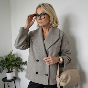 Puede incluir: Un abrigo gris claro de doble botonadura con grandes botones negros. El abrigo tiene una solapa ancha y se combina con un bolso beige. La persona lleva gafas con montura negra y un collar dorado. El estilo general es clásico y sofisticado.