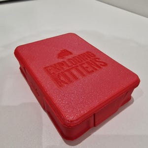 May include: A red rectangular box with the words "EXPLODING KITTENS" embossed on the top. The box has a textured surface and a small illustration of a kitten. The game's title is prominently displayed.