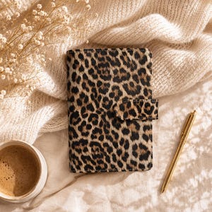 Puede incluir: Un cuaderno rectangular con un estampado de leopardo en tonos marrones y negros, que incluye una lengüeta de cierre magnético a juego. El cuaderno se encuentra junto a un bolígrafo de metal dorado, una taza de cerámica blanca con café y flores secas.