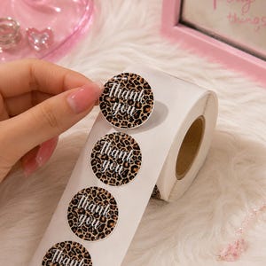May include: A roll of circular adhesive labels featuring a brown and black leopard print pattern. Each sticker displays the text Thank you in a white cursive script. The labels are arranged in a vertical line on a white backing paper roll.