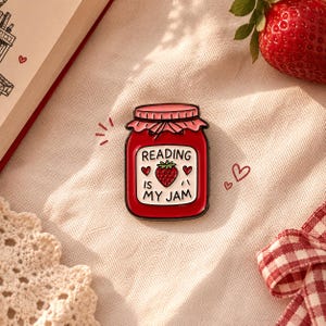 May include: A metal enamel pin shaped like a red strawberry jam jar with a pink lid. The white label features a small strawberry illustration, two red hearts, and the black text READING IS MY JAM. The pin has a black metal outline.