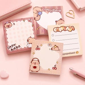May include: Four square sticky note pads featuring capybara illustrations. One pad has a heart-shaped writing area with the text capybara. Another has a checklist with the text capybara and keep a good mood. All pads feature pink and cream tones.