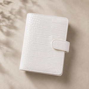 May include: A white personal planner with a textured crocodile skin pattern. The rectangular organizer features a secure strap closure on the right side and is constructed with clean, stitched edges throughout the entire exterior cover.