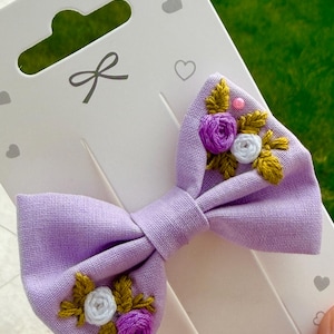 Peut inclure: Un nœud papillon pour cheveux en tissu lavande avec des détails floraux brodés. Le nœud présente un nœud central et est orné de fleurs brodées violettes et blanches avec des feuilles dorées. Le nœud est fixé à une carte blanche avec un motif de nœud.