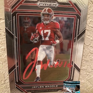 Puede incluir: Una tarjeta coleccionable Panini Prizm Draft Picks con Jaylen Waddle con el uniforme de la Universidad de Alabama Crimson Tide. La tarjeta tiene un borde plateado con detalles en rojo y negro. La firma del jugador es visible. El texto "JAYLEN WADDLE" y "THE UNIVERSITY OF ALABAMA" están impresos.