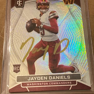 May include: A Jayden Daniels Washington Commanders rookie trading card featuring a holographic silver background with a radial pattern. The card displays a gold ink autograph, a Rookie RC logo, and the player's name printed at the bottom.