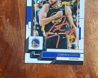 May include: A Donruss basketball trading card featuring Stephen Curry in a black Golden State Warriors jersey. The card displays a photograph of the player, the Golden State Warriors logo, and a brown ink autograph across the center. The text reads Stephen Curry and Warriors.