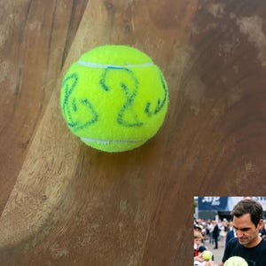 Può includere: Una pallina da tennis giallo brillante con il numero "2" e una linea curva disegnata con inchiostro blu. La pallina è appoggiata su una superficie di legno. L'immagine include anche una foto più piccola di una persona che firma una pallina da tennis simile.