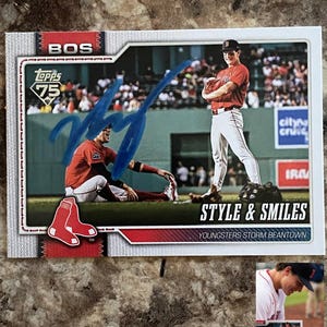 May include: A baseball card featuring two players in red and white uniforms. The card has the text "BOS" and "STYLE & SMILES". The Topps logo is visible. The card is signed in blue marker.