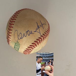 May include: A baseball with red stitching and a signature in blue ink. The baseball is a light tan color. A smaller image shows a person signing a baseball.