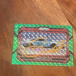 Könnte beinhalten: Eine Panini Prizm Sammelkarte mit einem hellblauen Rennwagen und einem Autogramm in goldener Tinte in der Mitte. Die Karte hat einen grünen Rand mit Karomuster, ein Prizm-Logo oben und den aufgedruckten Namen Bubba Wallace am unteren Rand.