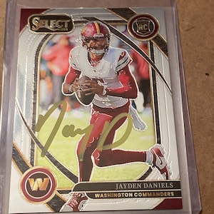 May include: A 2024 Select football trading card featuring Jayden Daniels of the Washington Commanders in a white and burgundy uniform. The card includes a gold ink autograph, a rookie card logo, and the team logo in a protective plastic sleeve.