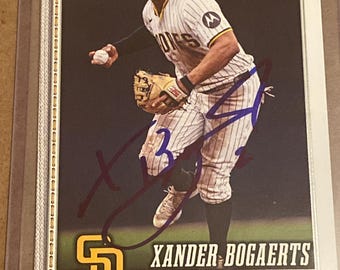 May include: A Topps 75 baseball card featuring Xander Bogaerts in a white pinstriped San Diego Padres uniform, holding a baseball and wearing a glove. The card includes the text SD, Topps 75, Xander Bogaerts, and Shortstop, with a dark ink autograph.