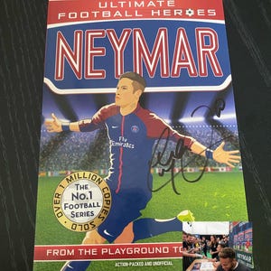 Pode incluir: Capa do livro "Ultimate Football Heroes Neymar" com uma ilustração de Neymar em uniforme de futebol. A capa é azul, vermelha e branca, com o nome do jogador em destaque. Inclui uma foto menor de Neymar autografando o livro.