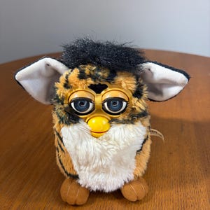 May include: A plush toy featuring a tiger-striped pattern in orange and black with a white chest and belly. It has large blue eyes, a yellow beak, black hair on top, and two large ears with white inner linings. The toy has two brown feet.