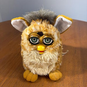 May include: A plush toy with golden-brown fur, a tuft of dark hair on top, and large green eyes with spiral patterns. It features prominent ears with white inner lining, a yellow beak, and two small feet. The toy has a soft, textured appearance.