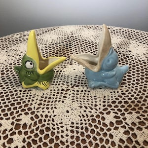 Puede incluir: Dos macetas de cerámica con forma de dinosaurio. Una es verde y amarilla, la otra azul y beige. Ambas tienen la boca abierta y están sobre un tapete de ganchillo. Las macetas son probablemente para pequeñas suculentas o plantas aéreas.