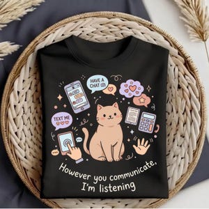 Puede incluir: Camiseta negra con una ilustración de gato de dibujos animados rodeada de bocadillos y gráficos de teléfono. El texto dice "Have a Chat", "Text Me" y "However you communicate, I'm listening". El diseño incluye corazones y estrellas.