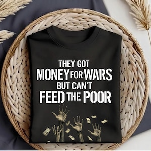 Op de afbeelding: Een zwart t-shirt met de witte tekst "THEY GOT MONEY FOR WARS BUT CAN'T FEED THE POOR" boven een illustratie van verschillende reikende handen en zwevende bankbiljetten. Het t-shirt heeft een ronde hals en korte mouwen.