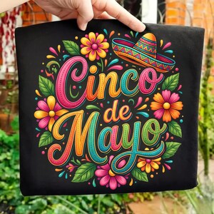 Puede incluir: Camiseta negra con un diseño colorido de Cinco de Mayo. El diseño presenta el texto "Cinco de Mayo" en letras vibrantes y multicolores, rodeado de flores y un sombrero.