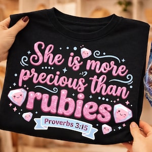 Puede incluir: Camiseta negra con la frase rosa "She is more precious than rubies" y dibujos de diamantes. También aparece el versículo bíblico Proverbios 3:15.