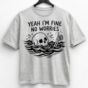 Puede incluir: Camiseta gris claro con un gráfico en blanco y negro de una calavera emergiendo del agua, con una mano de esqueleto haciendo una señal de aprobación. El texto "YEAH I'M FINE NO WORRIES" está encima del gráfico.