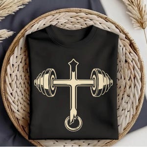 May include: Black crew neck t-shirt featuring a cream-colored graphic of a cross with a barbell design. The cross has a pointed top and a circular design at the bottom. The barbell design is on each side of the cross.