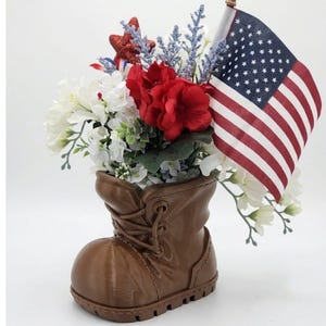May include: A brown boot-shaped vase filled with white and red flowers, blue sprigs, and a small American flag. A red glitter star adds a patriotic touch to the floral arrangement.