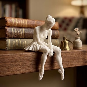 May include: A white matte figurine of a seated ballerina wearing a leotard and tutu, with hair in a bun and pointe shoes. The figure is positioned on the edge of a wooden surface, with three stacked books behind it, one labeled Sculpture and two Dance History.