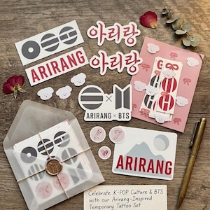 Peut inclure: Ensemble d'autocollants et de tatouages temporaires avec des textes et des motifs coréens. Les autocollants présentent le mot "ARIRANG" en rouge et blanc, ainsi que d'autres graphiques. Une petite carte indique "ARIRANG x BTS". Un stylo et une petite carte avec du texte sont également inclus.