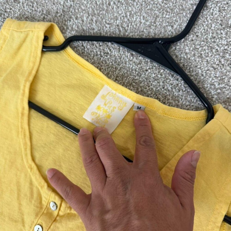 K&ouml;nnte beinhalten: Ein leuchtend gelbes Henley-Shirt mit V-Ausschnitt und drei wei&szlig;en Kn&ouml;pfen. Das Shirt h&auml;ngt an einem schwarzen Kleiderb&uuml;gel. Ein Etikett mit Text ist in der N&auml;he des Kragens sichtbar. Das Shirt ist Gr&ouml;&szlig;e M.