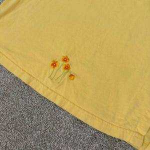 K&ouml;nnte beinhalten: Ein gelbes T-Shirt mit gestickten orangefarbenen und gelben Blumen. Die Blumen haben gr&uuml;ne Stiele und sind am unteren Rand des T-Shirts angeordnet. Das T-Shirt ist aus einem weichen, leichten Stoff gefertigt.