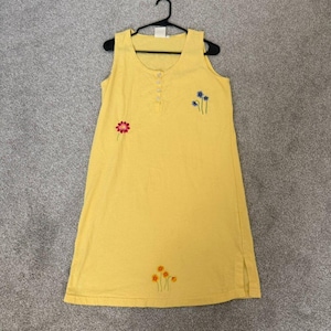 K&ouml;nnte beinhalten: Gelbes &auml;rmelloses Kleid mit gestickten floralen Details. Das Kleid hat einen Rundhalsausschnitt, eine Knopfleiste vorne und einen Seitenschlitz. Das Kleid h&auml;ngt an einem schwarzen Kleiderb&uuml;gel.